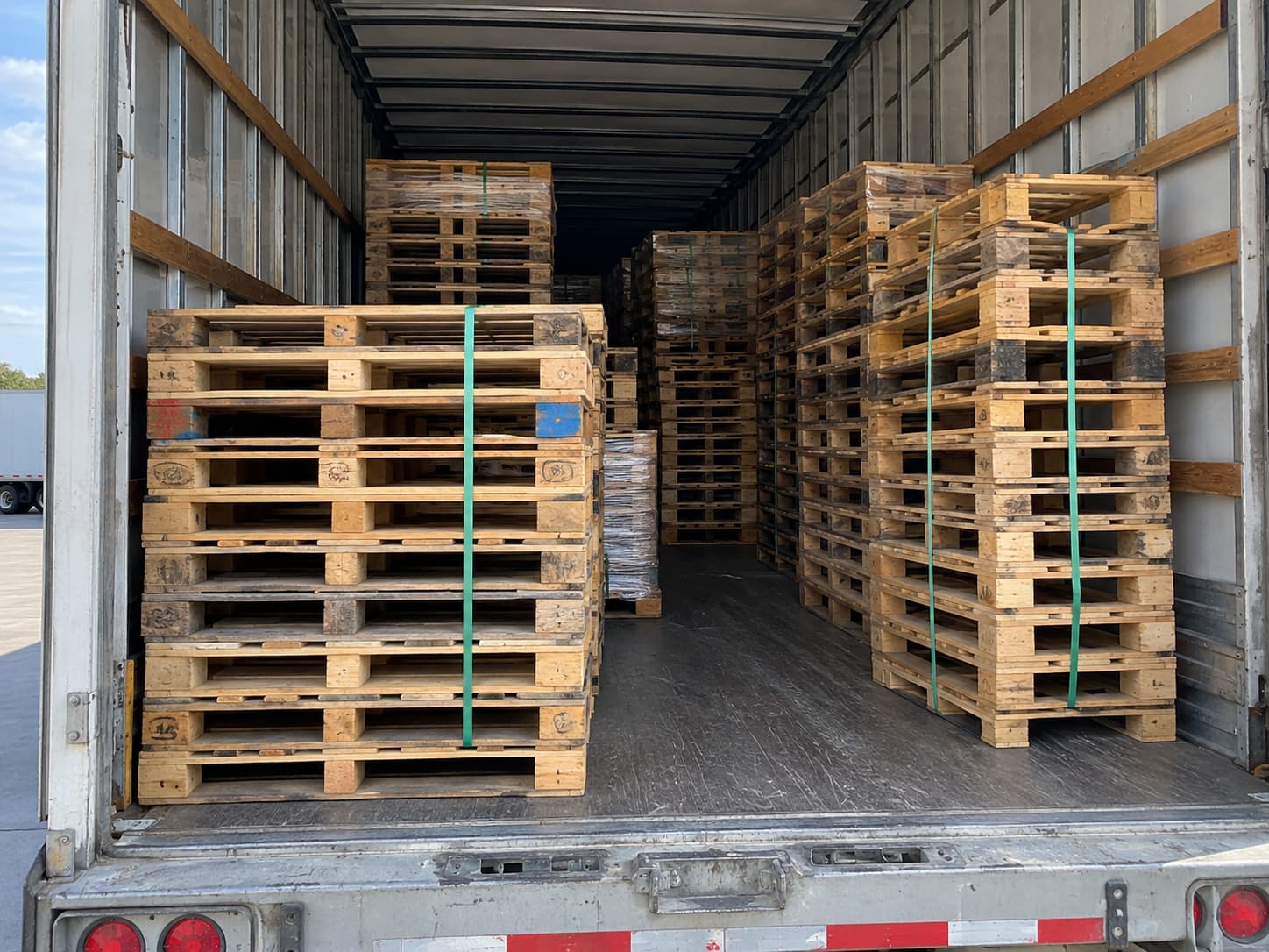 The open rear of a trailer loaded with banded stacks of wooden pallets, strapped in green banding.